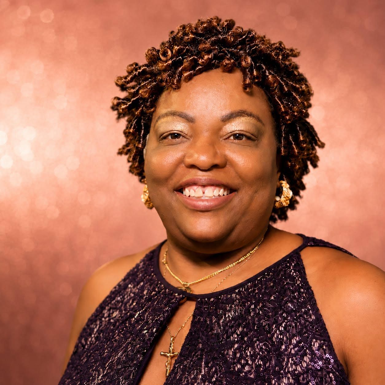 Woman smiling in front of a glittery pink background, wearing a sleeveless purple lace dress, gold earrings, and a gold necklace with a cross pendant.